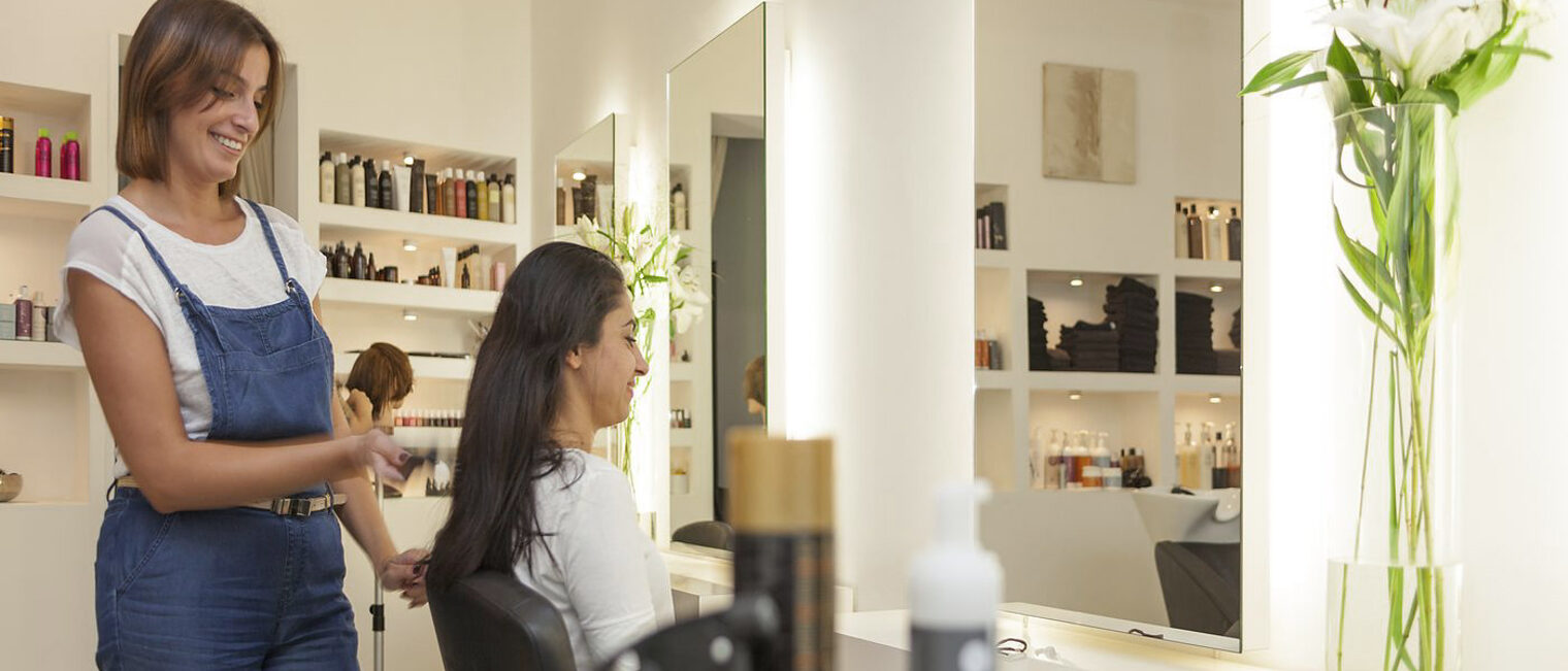 Friseurin in Latzhose Friseur, Friseurinnen, Friseurin, Friseurladen, Laden, Haare, Haare schneiden, Spiegel, Stuhl, Frau, Frauen, Blick in die Kamera, Latzhose, Flaschen, Dosen, Haarspray, Shampoo,
